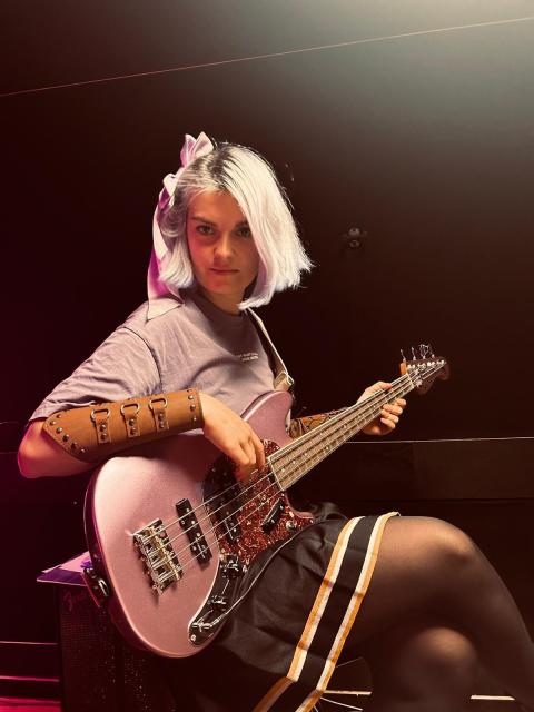A girl sitting on a barstool holding a lilac bass guitar on her lap. Her hair and Tshirt are also lilac, and she has a lilac bow in her hair. She's wearing a leather cuff on her arm and a pleated skirt. She's looking at the camera. 