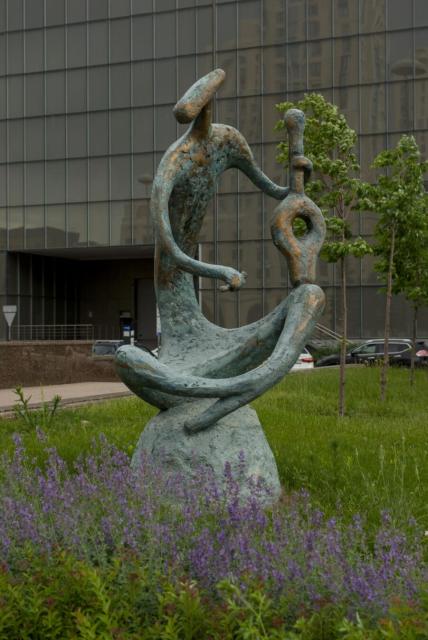The image features a bronze statue of highly stylised figure seated on a rock. The figure has an elongated head, thin torso ant thin long arms and legs. It's seated with its legs crossed with an amphora-like object that supposed to be a kobyz in its left hand and a short piece of what supposed to be a bow in its right hand. All the sculpture resembles some kind of ancient petroglyph.

The statue is situated outdoors and surrounded by purple flowers. In the background, there are some young trees and the glass wall of a building visible.