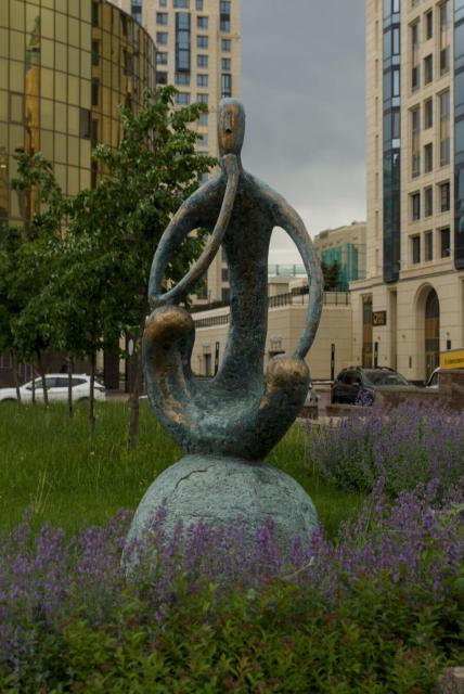 The image features a heavily stylised bronze statue of a person with thin torso and thin long arms seated cross-legged on a rock in the center. The right hand of the statue is bent with its elbow placed on its knee and its palm near its face. The left arm is rested on its knee. All the sculpture resembles some kind of ancient petroglyph.

The statue is surrounded by lush green plants and flowers, creating a serene atmosphere around it. In the background, there are tall buildings that add an urban touch to the scene. Cars can be seen parked on the street nearby.