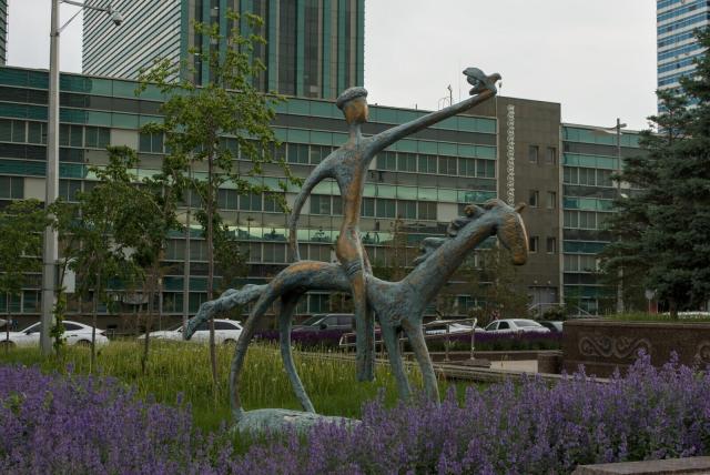The image depicts a serene park scene with a heavily stylised statue of a man riding a horse. Both figures have long thin limbs and thin torsos. A man has a bird on his left hand raised in the air. All the sculpture resembles some kind of ancient petroglyph.

The statue is surrounded by lush green trees and purple flowers, creating a harmonious blend of nature and art. In the background, there are tall buildings that add an urban touch to the otherwise natural setting.