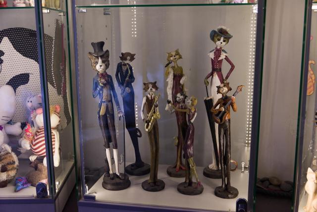 The image presents a collection of eight anthropomirphised cat figurines, each unique in its design and color. They are arranged on a glass display case that is part of a larger glass cabinet. The figurines include cats dressed in top hats and tails, cats dressed in lond dresses with sophisticateed hats, cats with saxophotes, cat with guitar, cat wit microphone and a pait of dancing cats. Each figurine has a distinct style and color with common idea of old jazz dancing, creating an interesting visual contrast against the transparent background of the glass display case.

The glass cabinet itself is quite large, occupying most of the frame from left to right. The arrangement of the figurines on this expansive surface makes them stand out prominently in the image.
