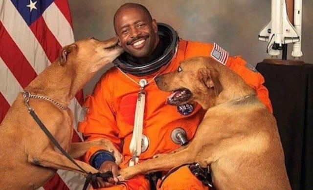 Astronaut Leland Melvin posing in his flight suit with his two dogs. He is smiling and looking at the camera, while the dogs climb on his lab and lick the side of his face. The flight suit is baggy and orange, with a black ring around the neck where the helmet attaches. A US flag is in the background, and a model of the space shuttle is partially visible.