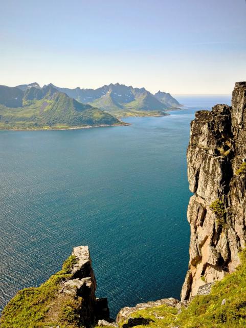 The image depicts a breathtaking coastal landscape with a clear blue sky. In the foreground, there are rugged cliffs with patches of green vegetation, including moss and small shrubs. The cliffs are steep and jagged, with a prominent vertical rock face on the right side. The middle ground features a calm, deep blue sea that stretches towards the horizon, reflecting the sky. In the background, a series of green, mountainous islands with sharp peaks rise from the water, some with snow-capped summits. The overall scene is serene and expansive, showcasing the natural beauty of the coastal region.