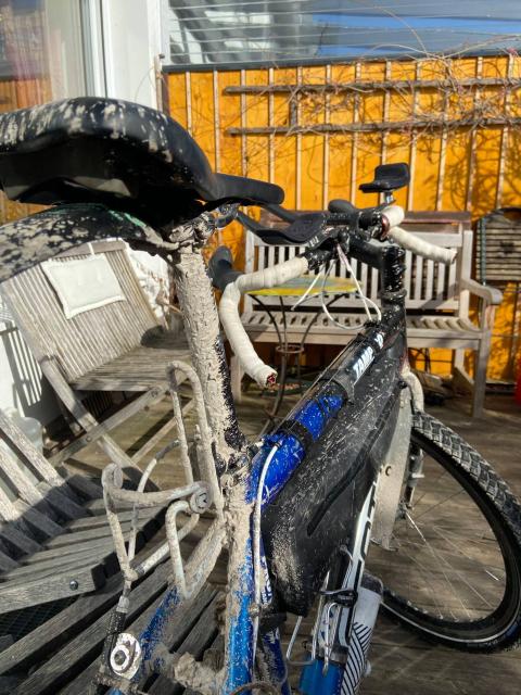 partial picture of a bicycle from the back. Showing seat, seatpost, top tube, bar and front wheel. The bike is dirty, a dried layer of fine mud covers all surfaces exposed to the wheels.
