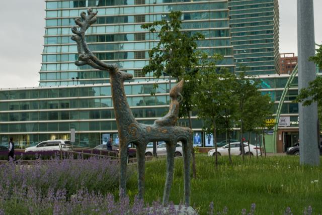 The image depicts a serene park scene with heavily stylized bronze deer statue with a smaller humanoid child-like figure sitting on its back. The deer has long thin legs and long thin antlers, its head turned back towards the child. All the sculpture resembles some kind of ancient petroglyph.

The sculpture is positioned in front of a modern glass building that reflects the surrounding environment. The park is lush with green grass and purple flowers, creating a picturesque setting for the statues. A few people can be seen walking around the park, adding life to the scene.