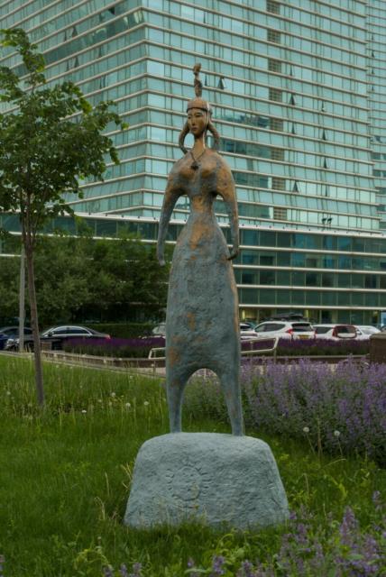 The image features a heavily stylized bronze statue of a woman standing on a rock in the center. The statue is adorned with a headdress and has her arms lowered to her hips. Apparently the woman wears knee-length dress and necklace with small pendant. She appears to be gazing towards the left side of the frame. All the sculpture resembles some kind of ancient petroglyph.

The statue is situated in front of a modern glass building that reflects the surrounding environment. In the background, there are several cars parked on the street. A tree can also be seen near the left edge of the image.