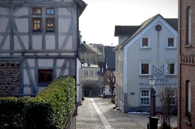 A view into a narrow cobblestone road going downhill on a sunny February day. To the left a half-timbered house, to the right other historic houses. One shows an inscription for a stoneware fabric. Old lanterns are visible. In the background the roads seems to disappear in a right turn and above some more older houses become visible.