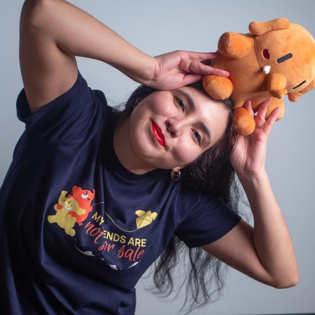 A beautiful woman with dark hair, gold earrings and red lipstick, wearing a french navy colored t-shirt with a colorful screen printed design on the front, while holding a red Plushtodon on her head.