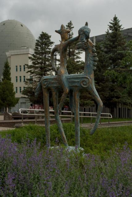 The image depicts a bronze statue of two heavily stylised figures. One figure is a horse with long thin legs and almost straight horizontal body and the other figure is a man sitting on a horse wearing a sophisticated hat resembling mongolian and holding some kind of a predator bird on his straight right hand. The horse is depicted in a dynamic pose with one of its forefoot in the air and its head turned to its back looking at the man.

The statue is situated in a park setting, surrounded by lush green trees and purple flowers. In the background, there are buildings visible behind the foliage.
