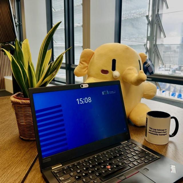 A laptop with the lock screen showing a Commission logo and the time 15:08. Behind the laptop on the desk sits the mastodon mascot, a little yellow elephant. There is also a coffee cup, and a plant on the table. In the background, you can see the iconic window structures of the European commission headquarter the Berlaymont building. And you can just about make EU flags below on the street