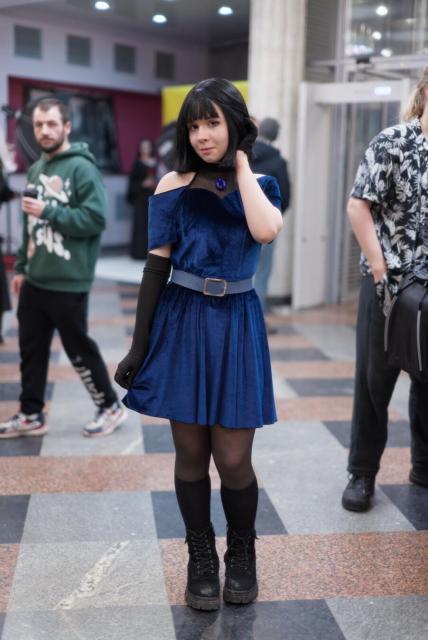 The image shows a person standing in a public indoor space. The person is wearing a blue, short, off-the-shoulder velvet dress with short sleeves. Across the chest is a dark mesh panel featuring a large blue gem at its center, and a light blue-gray belt with a metallic buckle is around the waist. They have black elbow-length glove on the right hand and short black glove on the left, black thigh-high stockings, and black lace-up combat boots. Their hair is black, shoulder-length, and has bangs. Their right hand is down holding a hem of their skirt, their left hand is near their neck as if they arrange their hair.

In the background, the indoor setting has a checkered floor with gray, brown, and black tiles. Other people are present, but out of focus. The space includes columns, doors, and ceiling lights providing illumination.