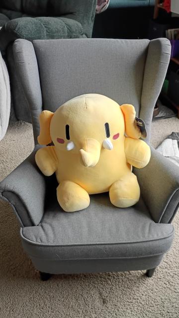 a small size chair with the large tan mastodon plushie, which is a cartoon elephant shaped critter with vertical dot eyes and oval blushes cheeks.  finally a cute trunk with tiny tusks.  the plushie comfortably fills the chair.