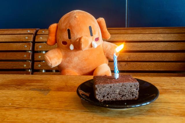 Plushtodon is sitting at a table in EPFL’s Arcadie cafeteria. A brownie with a candle in it is placed in front of him.