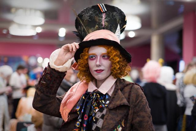 The image shows a person dressed in a costume that resembles the Mad Hatter from the "Alice in Wonderland" story. The individual is wearing a tall, black top hat adorned with colorful ribbons and a small feather. The outfit includes a brown jacket with a patterned scarf and a light-pink shirt with a collar. A white, ruffled sleeve is visible on one arm. Around their neck is a colorful scarf with patterns like circles. The person has curly red hair and is applying makeup, which includes white face paint and blue and pink eyeshadow. The background shows an indoor space with other people, pink walls, ceiling lights, and blurred figures, suggesting a crowded event or convention.