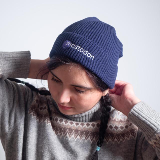 A beautiful woman with dark hair and two braids, wearing a grey wool sweater and a blue fisherman beanie with the Mastodon logo embroidered on the front.
