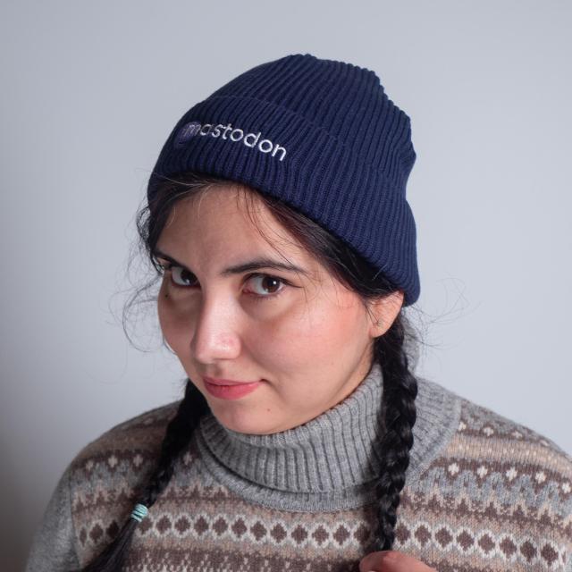 A beautiful woman with dark hair and two braids, wearing a grey wool sweater and a blue fisherman beanie with the Mastodon logo embroidered on the front.