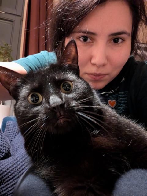 A selfie style photograph of a woman with a black cat sitting on her lap. They're both looking at the camera with interesting expressions.
