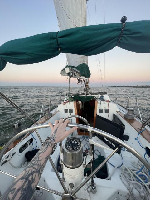 A view from the deck of a sailboat, showing a hand on the steering wheel. The boat is on water with a distant horizon, and the sky features a gradient of colors during sunset. Various sailing equipment and ropes are visible in the foreground.
