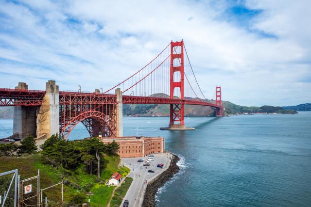 An orange bridge spans a gap between two land masses, with a comparatively-small old fort underneath. [Ricoh GR III]
