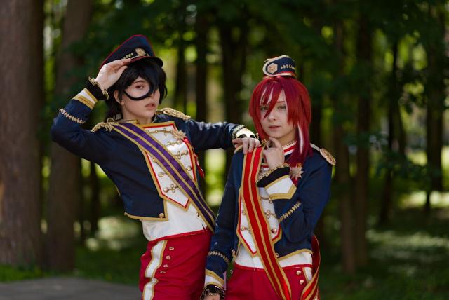 Two individuals stand outdoors in a wooded area with tall trees and blurred green foliage in the background. Both wear matching dark blue military-style uniforms: jackets with white and gold trim, red pants with white stripes, and red sashes with gold edges. The person on the left has a dark-blue cap with a gold emblem, a black hair lock is covering one eye; they salute with their right hand or adjusting their cap. The person on the right has red hair and a smaller cap resembling a garrison cap in the same style with a gold emblem, placing a hand near their neck. The uniforms feature gold buttons, decorative shoulder straps, and matching details on the sleeves. The scene is well-lit, with natural sunlight filtering through the trees.
