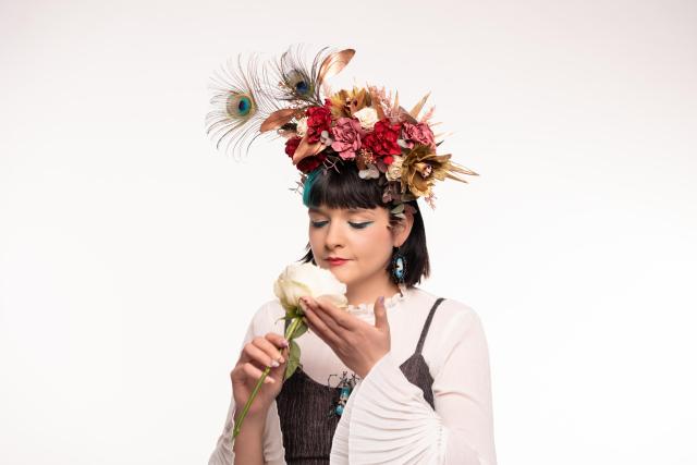 a studio photograph that I took of my friend and fellow artist Blumen Mitzi. She is wearing a crown of flowers and holding a white rose. she gentle holds the rose to her face as if inhaling its scent