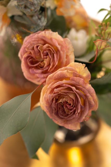 a close-up photo of two darkish off-pink roses between leaves and other blurry flowers, sticking out from a blurry golden vase