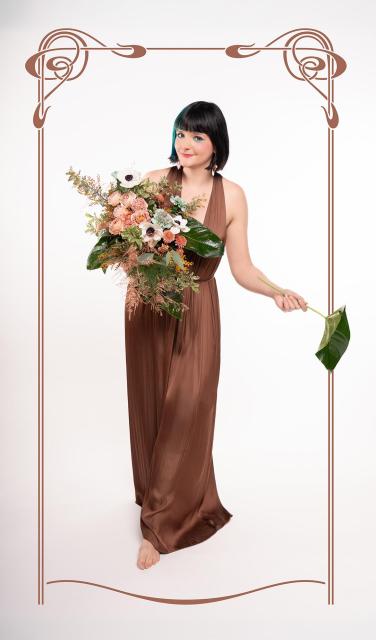 a studio photograph that I took of my friend and fellow artist Blumen Mitzi. She is holding a luscious bouquet of flowers while one of her arms is held out to the right, holding a leaf. the pose is reminiscent of images by art nouveau artist Mucha. a Jugendstil-like ornament is framing the image