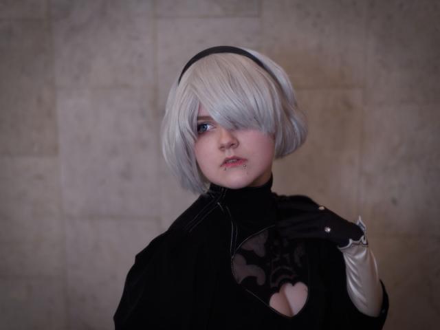 The image features a cosplayer dressed as 2B from the video game *NieR: Automata*. The cosplayer has short, choppy, light grey hair styled in a classic 2B cut. She has light blue eyes and a subtle pout. She's wearing a small, discreet lip piercing. She’s wearing 2B's iconic black turtleneck, which is detailed with a distinctive cut-out design resembling a stylized heart. Her sleeves are white and fitted, with a similar cut-out shape. She's wearing black gloves with matching cutouts. She’s looking to the left side of the frame with a serious, slightly melancholic expression. Her  left hand is raised to her chest.
The background is a textured, muted grey wall, providing a simple, neutral backdrop that allows the cosplayer's outfit and expression to stand out.
