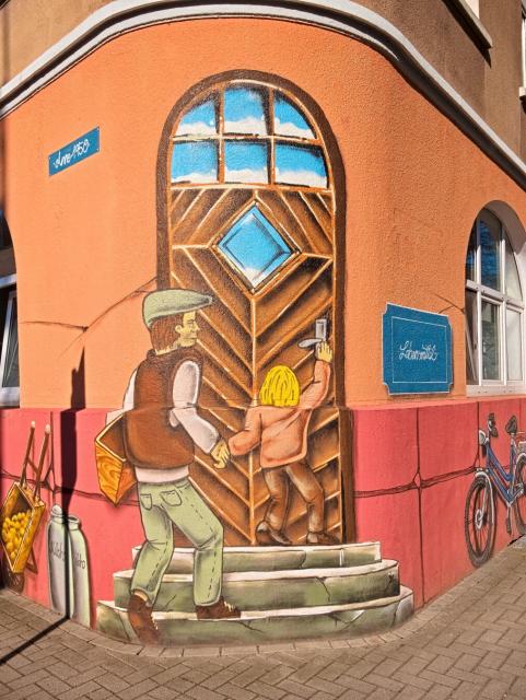 We see a painted house façade. It is orange, with its lower third painted red. Directly in front of us, on the rounded corner of the house, a door has been painted onto the house. It is brown with a small diamond-shaped window and smaller windows above the door. A concrete staircase has been painted below the door. We see a child and a man approaching the door. The child has its back to us and has raised its right arm to press down the door handle. The man is just stepping onto the first step of the staircase. He is wearing a flat cap and carrying a bag under his arm. On the right side of the house wall, there is a blue bicycle, and on the left, milk cans and a wheelbarrow with potatoes.
The picture is wonderfully colourful and drawn in great detail. It really puts me in a good mood.