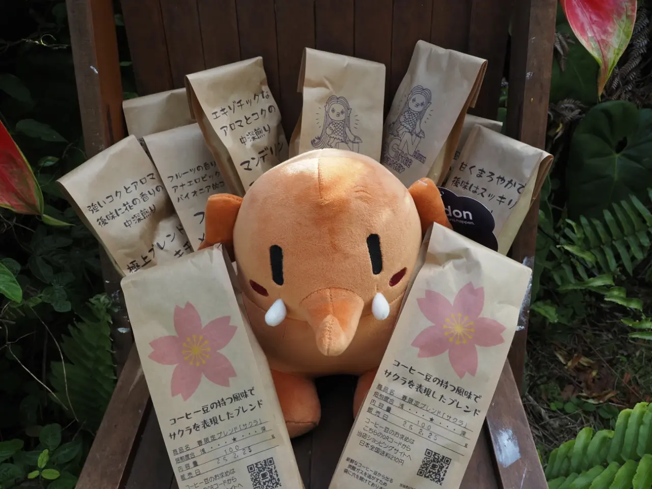 アンスリウムの前でコーヒー豆の袋に囲まれてポーズする小さいぞうさんのぬいぐるみの写真