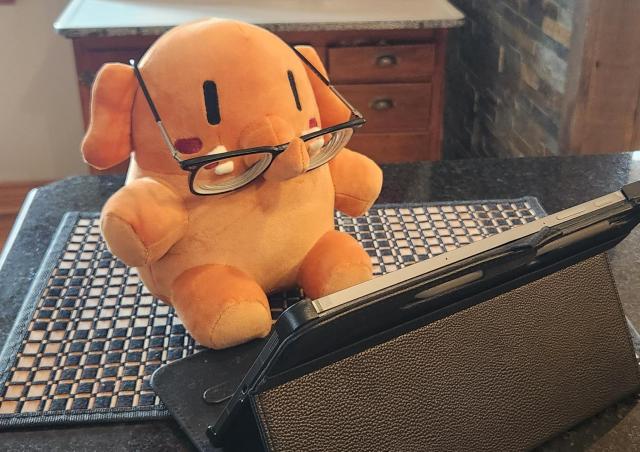 A peach coloured Plushtodon, wearing reading glasses,  sits in front of a tablet that is propped up for optimal viewing.