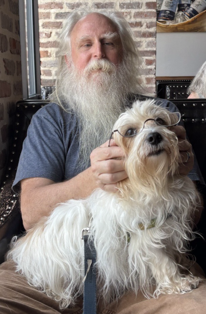A shaggy old man with a shaggy little dog on his lap. Long white hair and beard on both, tan highlights for the dog. Dog is wearing the man's half glasses.