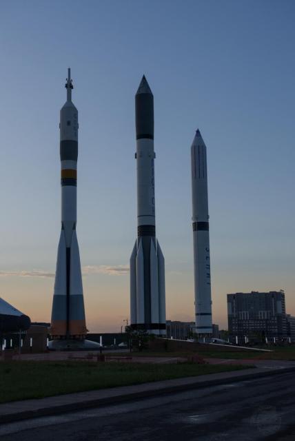 The image shows three large rockets displayed outdoors. The rockets are positioned side by side, with the one in the center being the tallest and the one on the right being the shortest. Each rocket has a distinct color scheme and design, with visible text and markings on their bodies. The leftmost one is “Soyuz”, in he middle is “Proton”, to the right is “Zenit”.

The background features a clear sky with a hint of sunset, and there are some buildings and structures visible in the distance. The setting appears to be a public display or museum exhibit.