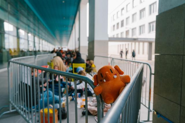 Plushtodon, lors de la mise en vente des billets pour Balélec. La peluche est posée sur une barrière, à la première place de la file d'attente. Au second plan, des personnes faisant la queue sous le couvert du bâtiment BM