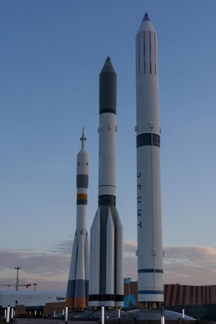 The image shows three large rockets displayed outdoors. The rockets are arranged in a row, diagonally from left (the farthest one) to right (the nearest one). on the left is “Soyuz”, in the center stands “Proton” (the largest of all), to the right is “Zenit”. The rockets are predominantly white with black, gray and orange sections. The nearest rocket has a distinctive purple and white tip. The background includes a clear blue sky with some clouds, and there are some structures and buildings visible in the distance. The setting appears to be a space museum situated near the Kazakhstan National Space Centre.
