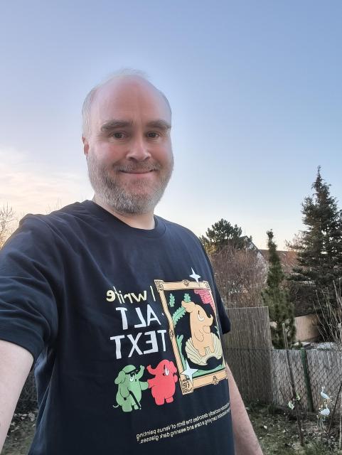A middle aged man showing off his new 'I write Alt Text' Mastodon shirt