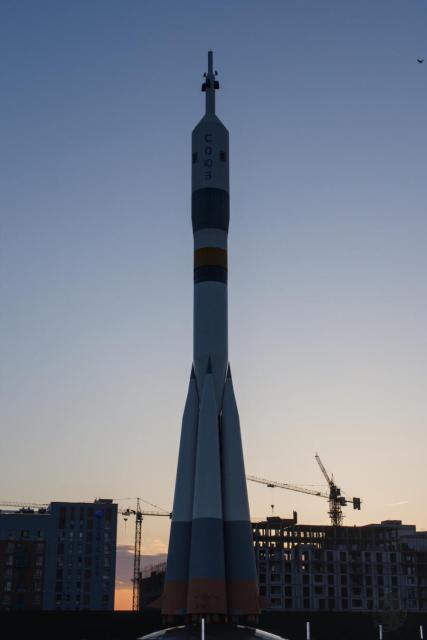 The image shows a large rocket model prominently displayed against a backdrop of a clear sky during what appears to be a sunset, as indicated by the warm hues near the horizon. The rocket is white with orange and gray sections, and it has the letters "SOYUZ" written vertically in Cyrillic script on its body, indicating it is a model of a Soyuz rocket. The rocket is positioned in an urban setting, with buildings and construction cranes visible in the background.