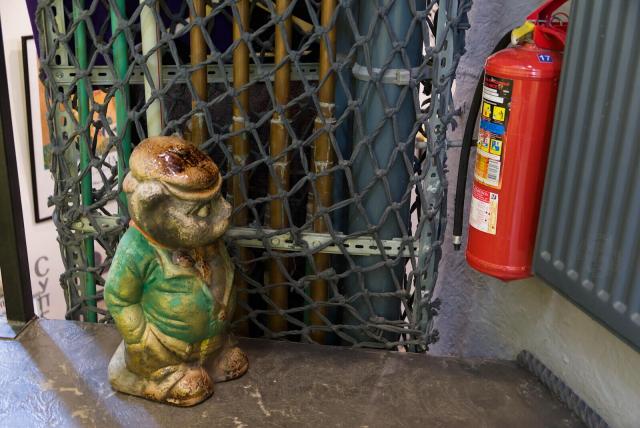 The image captures a quaint scene featuring a ceramic figurine of an anthropomorfic cat in a green suite positioned in an indoor setting. The figurine, which is placed in the left half of the frame and appears to be made of green terracotta with patches of yellow glaze on its head and chest, stands out prominently and is adorned with a brown hat.

To the right, slightly in the background but still noticeable due to its size, there is a red fire extinguisher hanging in the corner - an unexpected but striking contrast against the otherwise muted tones. The ceramic cat is positioned such as it looks at the fire extinguisher with a bit of distrust and surprise.

In the background rope net fences plastic pipes of different size. THey are mostly gray, green and olive.

The backdrop reveals a setting that appears both rustic yet functional. The floor is concrete, bearing signs of age with patches where paint has chipped off in varying shades of brown and beige.