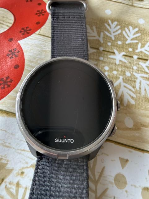 A close-up of a Suunto smartwatch with a black strap, placed on a decorated surface featuring red and white snowflake patterns. The watch face is dark and inactive.