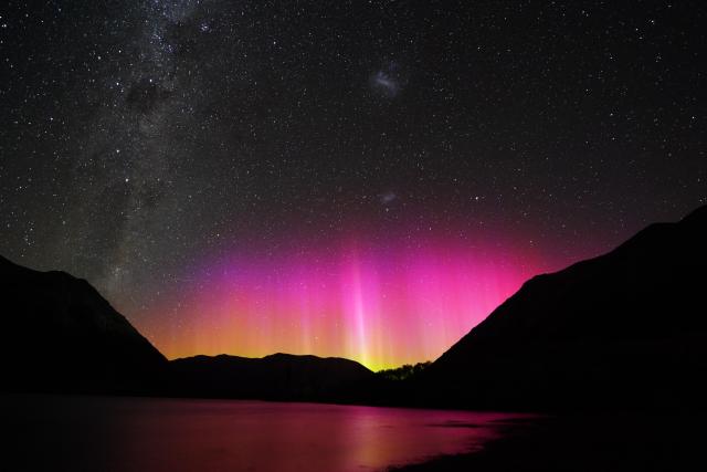 A nighttime view looking to the south, with the Milky Way stretching across the left portion of the photo, and the two Magellanic clouds visible as little puffs of cloudy stars, with blazingly bright pink and orange aurora pillars low on the horizon.  The auroras are also visible reflected in a lake that's just barely visible in the starlight, with silhouetted mountains on each side of the image.