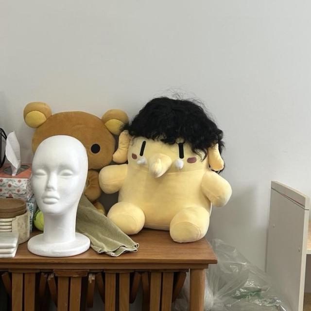 Plushtodon plush toy wearing a black hair wig, that makes him look like he's a fancy dapper guy (or Rihanna). He sits on a brown table next to a Rilakkuma plushie, and a Styrofoam head structure for the wig.