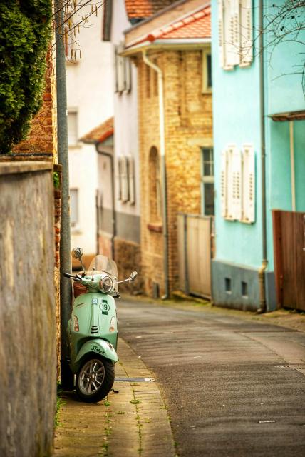 Eine hellgrüne Vespa ist im Vordergrund links in einer engen Gasse geparkt. Das historische Gebäude links hat eine Ziegelwand, während die Gebäude im Hintergrund verschiedenfarbig sind: eines ist gelb, ein anderes hellblau gestrichen und hat weiße Fensterläden. Die Gasse führt in die Ferne, und die Perspektive schafft Tiefe. Auf der Vorderseite des Rollers ist die Nummer "19" in einem Kreis zu sehen, und auf dem vorderen Kotflügel steht das Wort "Sister".