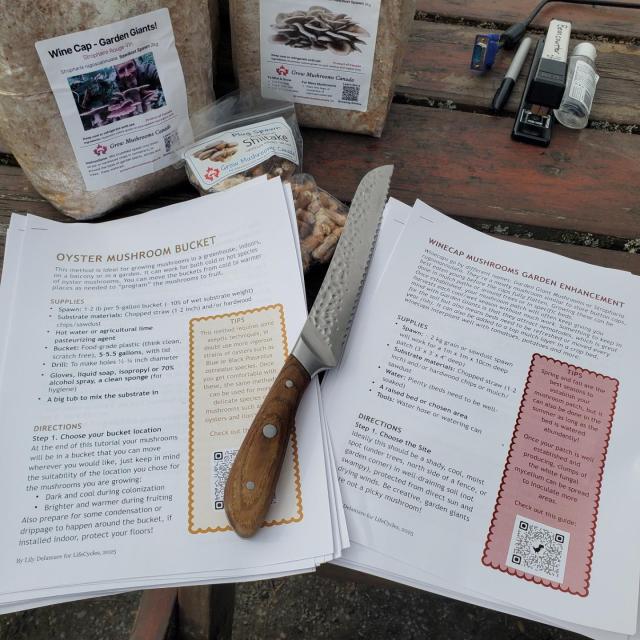 A close-up of a wooden table with several objects: a stainless steel knife with a wooden handle, two large plastic bags labeled "Wine Cap - Garden Giants!" and "Grow Mushrooms Canada," a bag of shiitake mushrooms, two staplers, a permanent marker, a computer cable, a bottle of hand sanitizer, and several stapled instruction sheets about growing mushrooms.