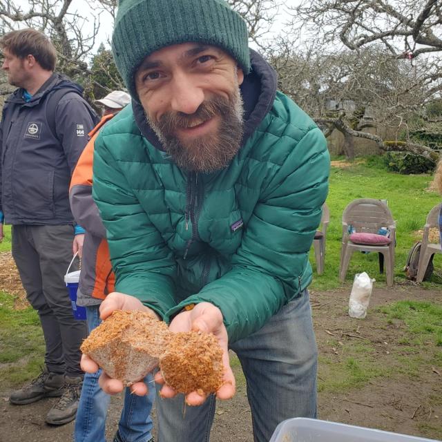 Man with a beard and green beanie and jacket smiles and holds two pieces of tan, porous material in his hands, with other people in the background.
