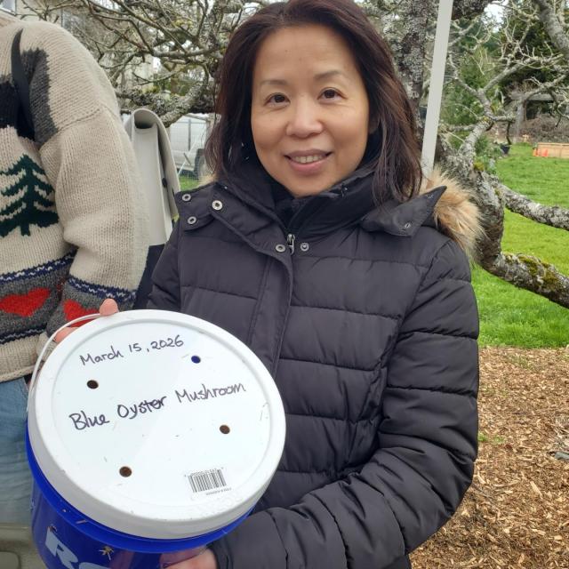 Smiling woman in a black puffer jacket holds a blue and white bucket labeled "Blue Oyster Mushroom, March 15, 2026", with a backdrop of a blurred outdoor scene.