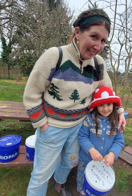 Woman and young girl posing for a photo outdoors. The woman is wearing a cream-colored sweater with a mountain scene design, blue jeans, and a braided headband. The girl wears a red mushroom-shaped hat and blue sweater, holding her blue oyster mushrooms grow bucket. They are standing in a community orchard park.