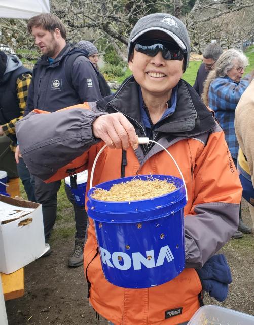 Smiling person wearing sunglasses and an orange jacket, holding a blue RONA bucket filled with straw, other people are in the background.