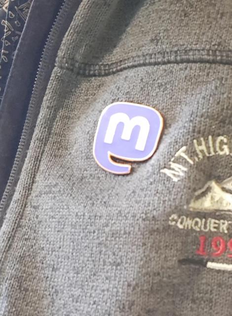 A shiny and official 3x3cm sized Mastodon enamel pin on my greyish sweater. It looks like an abstract Mastodon head with trunk and as speech bubble at the same time. What a great design combining these two aspects.