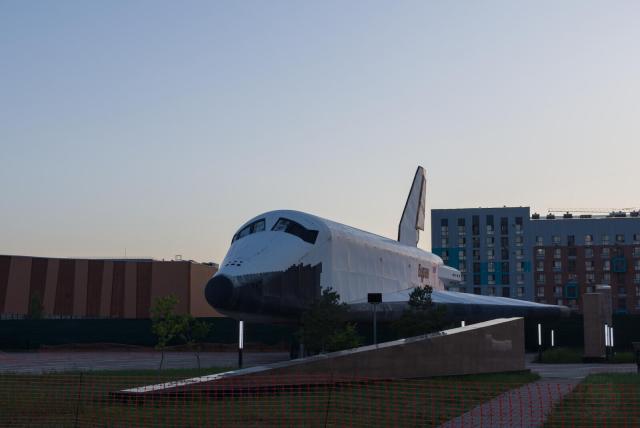 The image displays a space shuttle mounted on a display structure, prominently positioned against a backdrop of urban buildings during what appears to be twilight. The shuttle, a symbol of space exploration, is the central focus. The surrounding area is a mix of industrial and modern architectural elements, including a large brown building with vertical stripes and a range of colorful high-rise structures in the distance. The foreground features a red netted barrier, suggesting restricted access to the display area. The sky is clear with a gradient of colors near the horizon, indicating dusk.