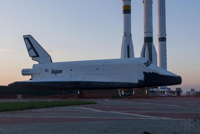 The image displays a large, white space shuttle with the word "Buran" written on its side, indicative of the name of the Soviet Union's first space shuttle. The shuttle is stationed on a paved area with a backdrop of multiple launch rockets, suggesting a space exhibition or some sort of museum. The setting appears to be during twilight, as the sky is illuminated with a soft light, hinting at dusk. The environment includes a mix of natural and urban elements, with visible grass and the distant silhouette of buildings, which could be part of a city or industrial complex. The shuttle and the rockets are the central focus, and their prominent positioning and lighting emphasize their significance.